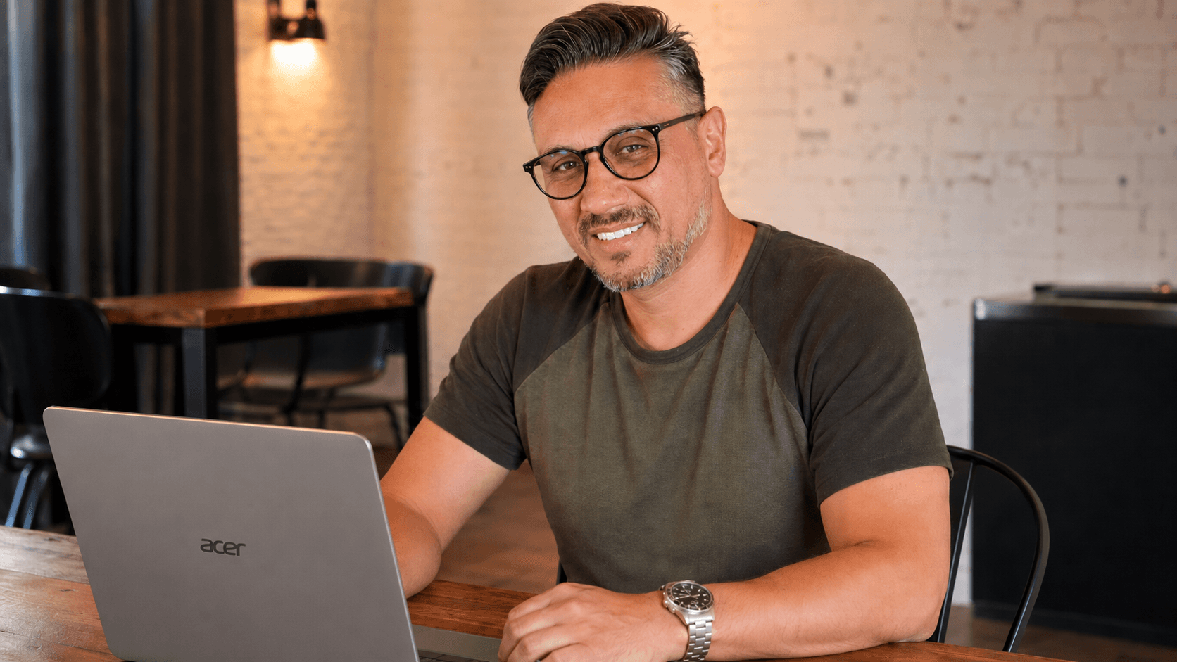 Hombre con gafas y barba sonríe frente a una computadora portátil Acer en una cafetería.